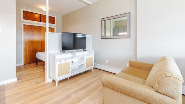 A living room with a white entertainment unit, a beige sofa, and a wooden door. - Cozy Studio w/ Full Kitchen (Hampton)