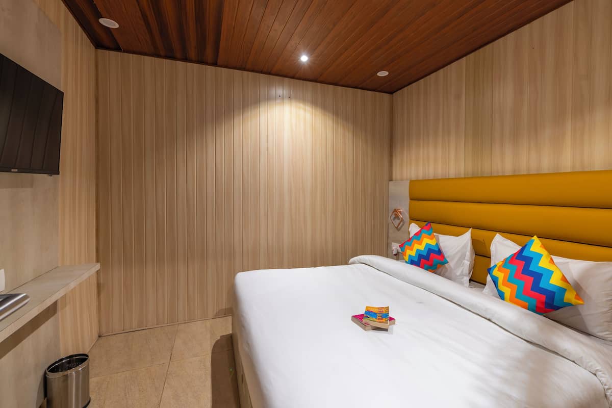 A neatly arranged hotel room with a wooden ceiling, a bed with white bedding and colorful zigzag pillows, and a small trash bin.