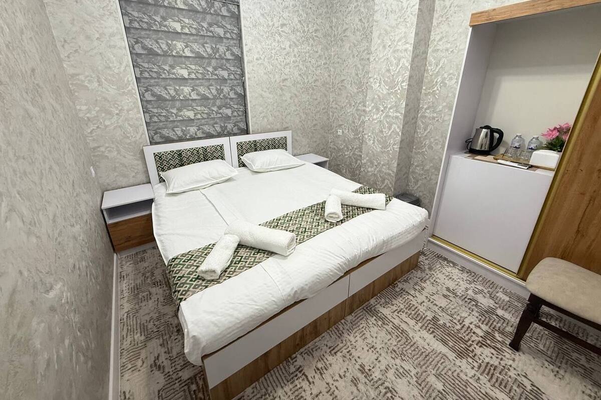 A neatly made bed with white linens and a patterned pillow, a bedside table with a kettle, and a chair in a room with patterned walls and a wooden cabinet.
