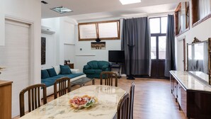 Ein geräumiges Wohnzimmer mit Essbereich, einer Couch, einem Fernseher und einem großen Fenster mit Vorhängen.