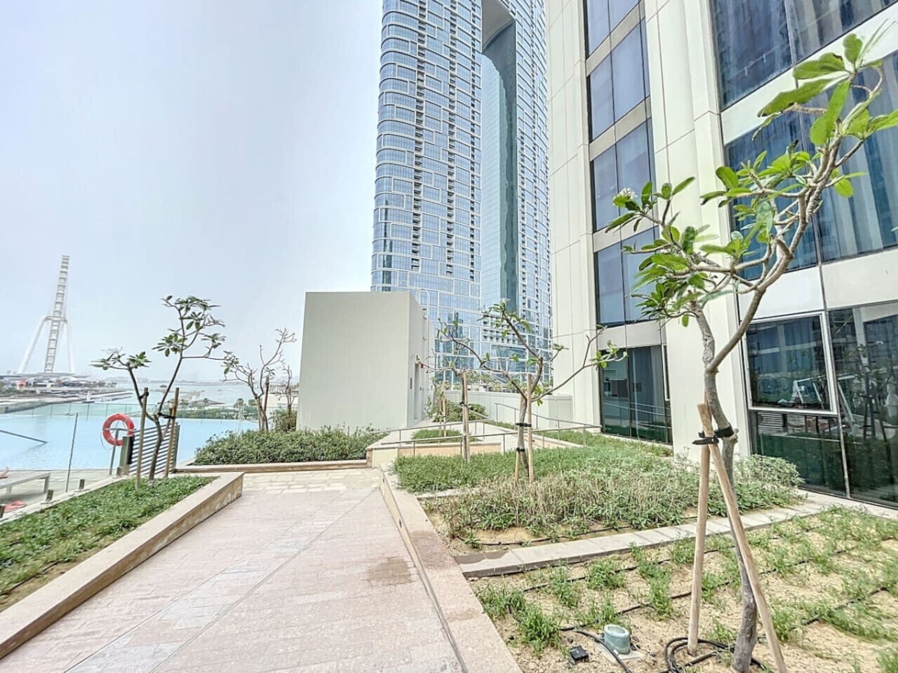 Un edificio moderno con fachada de vidrio, un área ajardinada con árboles jóvenes y una vista de un puente al fondo.