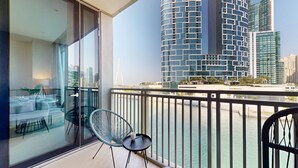 A balcony with a glass door, a round table, and two chairs. A cityscape with modern buildings and a bridge is visible in the background. - Elegant 1 Bed | Canal Views | Close to Ain Dubai I 5242 Tower 2 (Dubai Marina)