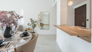 A modern dining area with a glass table, chairs, and a vase of flowers. There is a mirror on the wall and a wooden counter to the right.