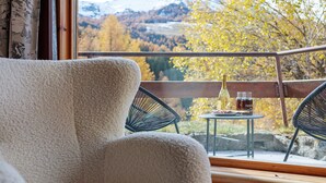 A cozy room with a plush chair, a small table with a bottle and glasses, and a scenic view of autumn foliage through a window.