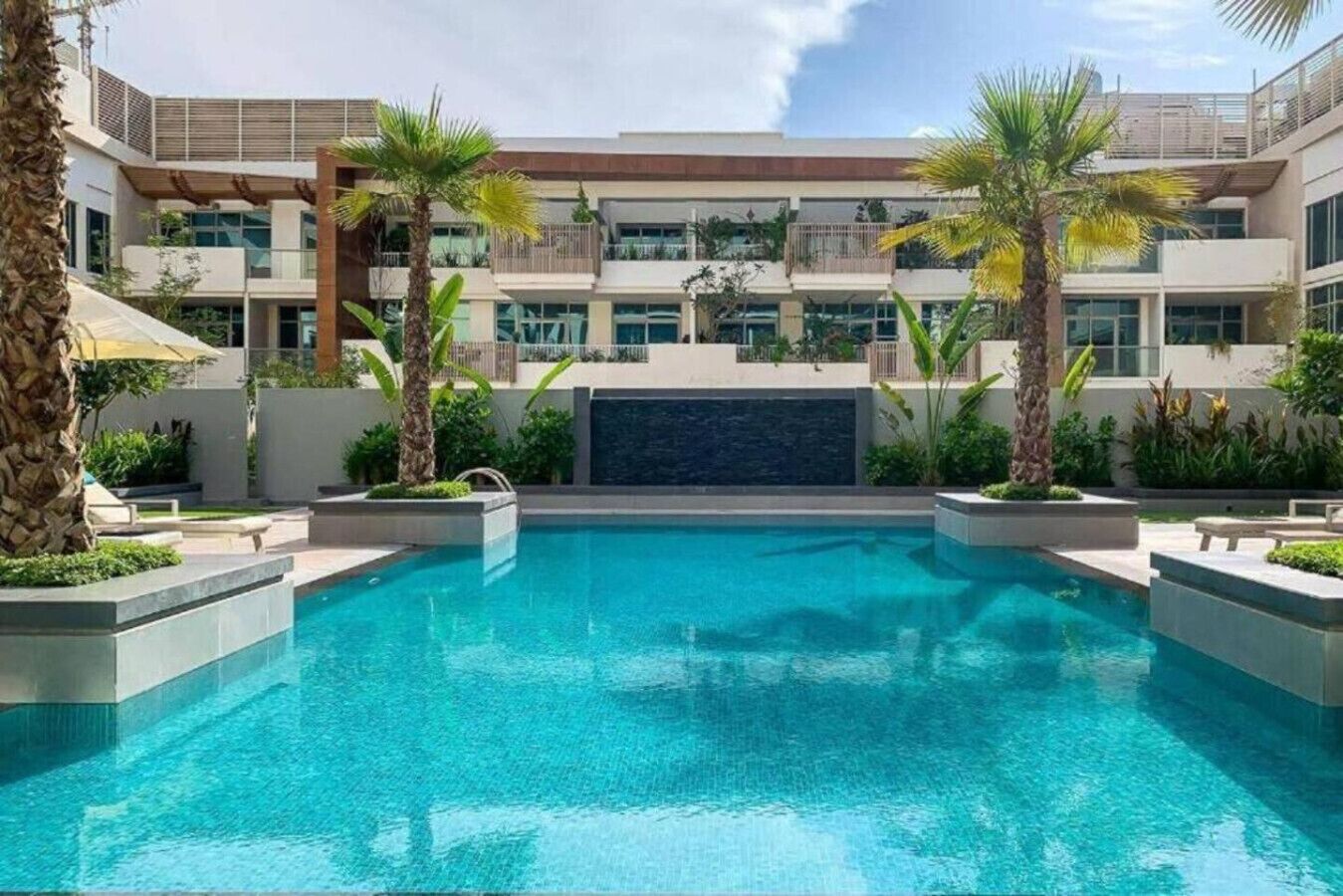 A modern hotel with a central swimming pool, surrounded by palm trees and a multi-story building with balconies.