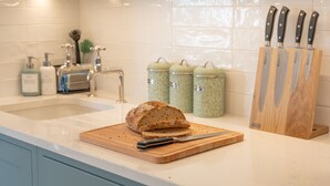A kitchen with a loaf of bread on a cutting board, a knife, and a knife block. There are two sinks, a faucet, and a soap dispenser. - The Little Stable - Cosy 1 Bed Cottage in Burford (Burford)