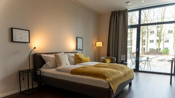 A modern hotel room with a large bed, a bedside table, a lamp, and a view of the outside. - Arbio I Duxen Apartments Hagenbeck Zoo (Hamburg)