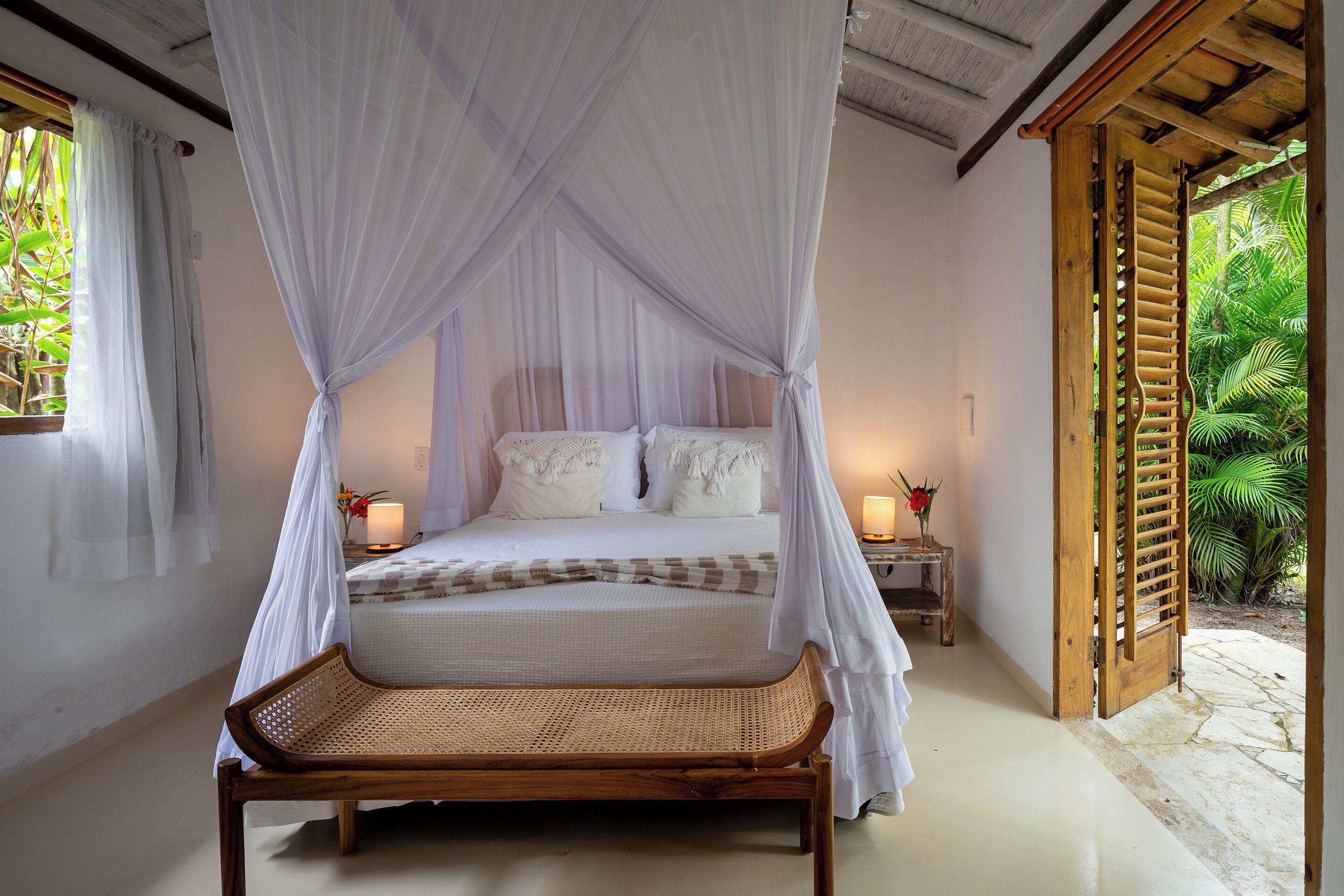 A room with a bed under a canopy, a wicker chair, a nightstand with a lamp, and a wooden door leading to a garden.