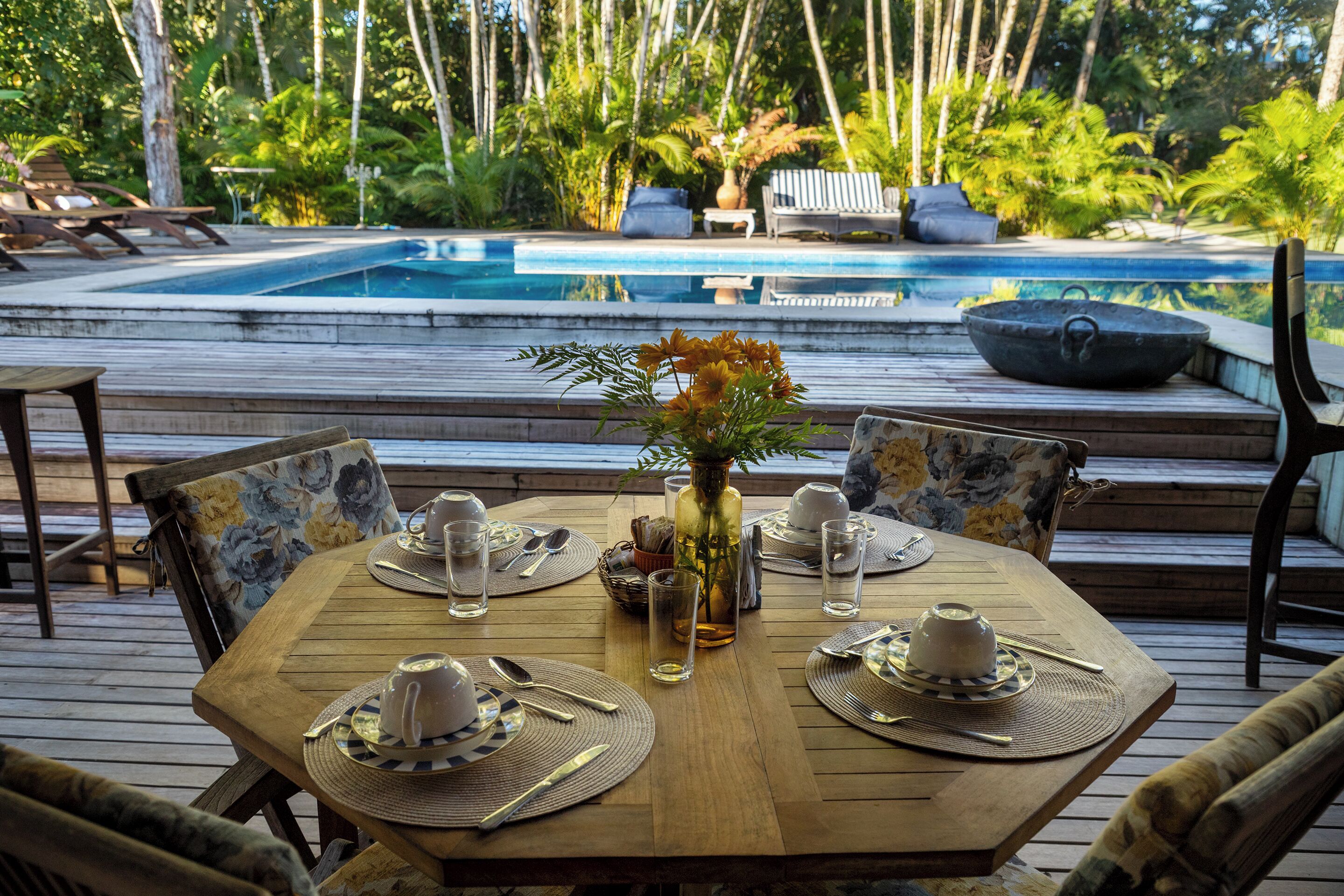 Terraza de madera con una mesa de comedor preparada para cuatro personas, rodeada de sillas, con vista a una piscina.