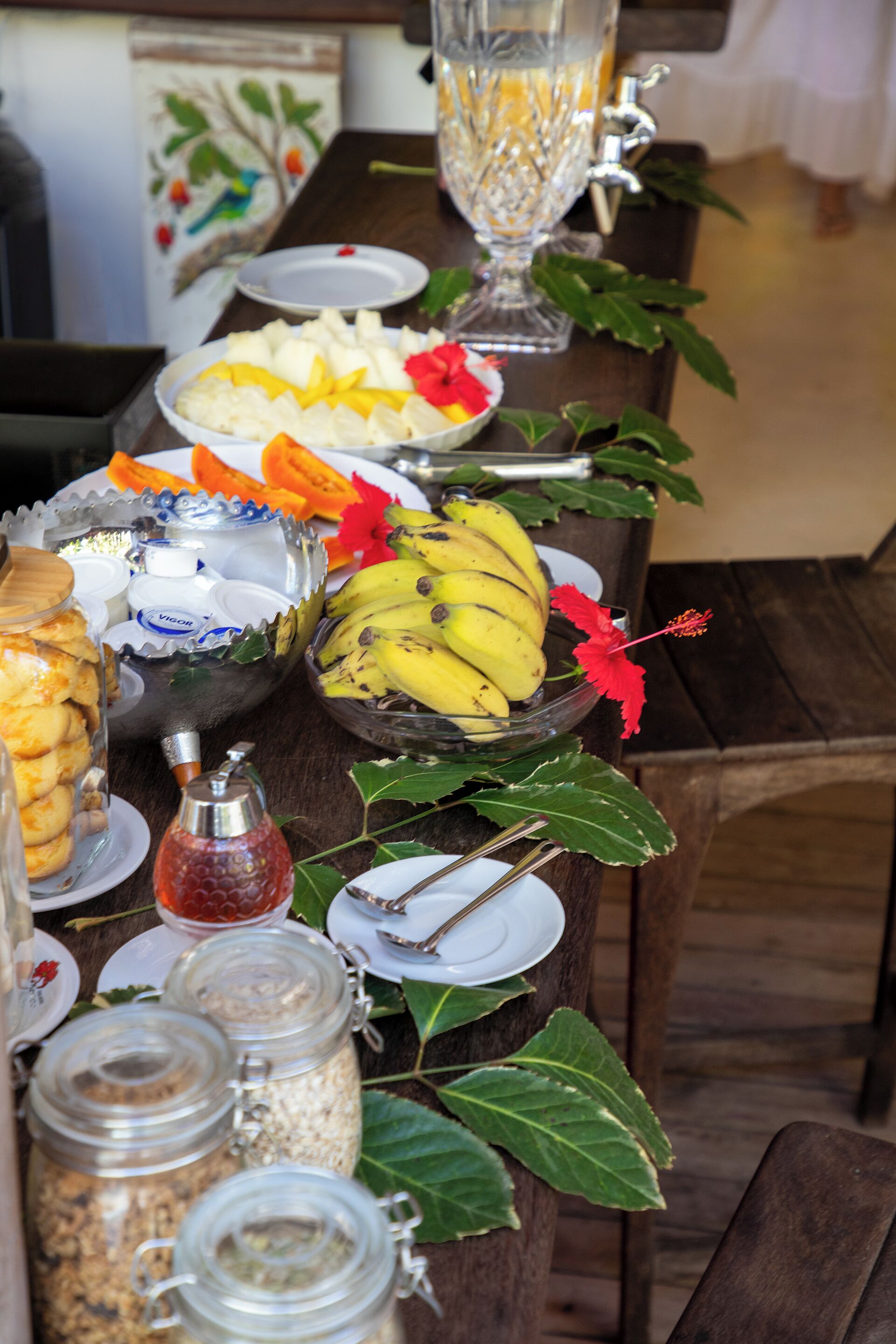 Une table sur laquelle on trouve divers aliments et condiments, notamment des bananes, un saladier de fruits et des pots de confitures.