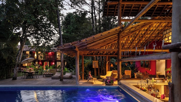 A pool with a wooden deck and lounge chairs, surrounded by lush greenery. - Pousada Hibisco (Porto Seguro)