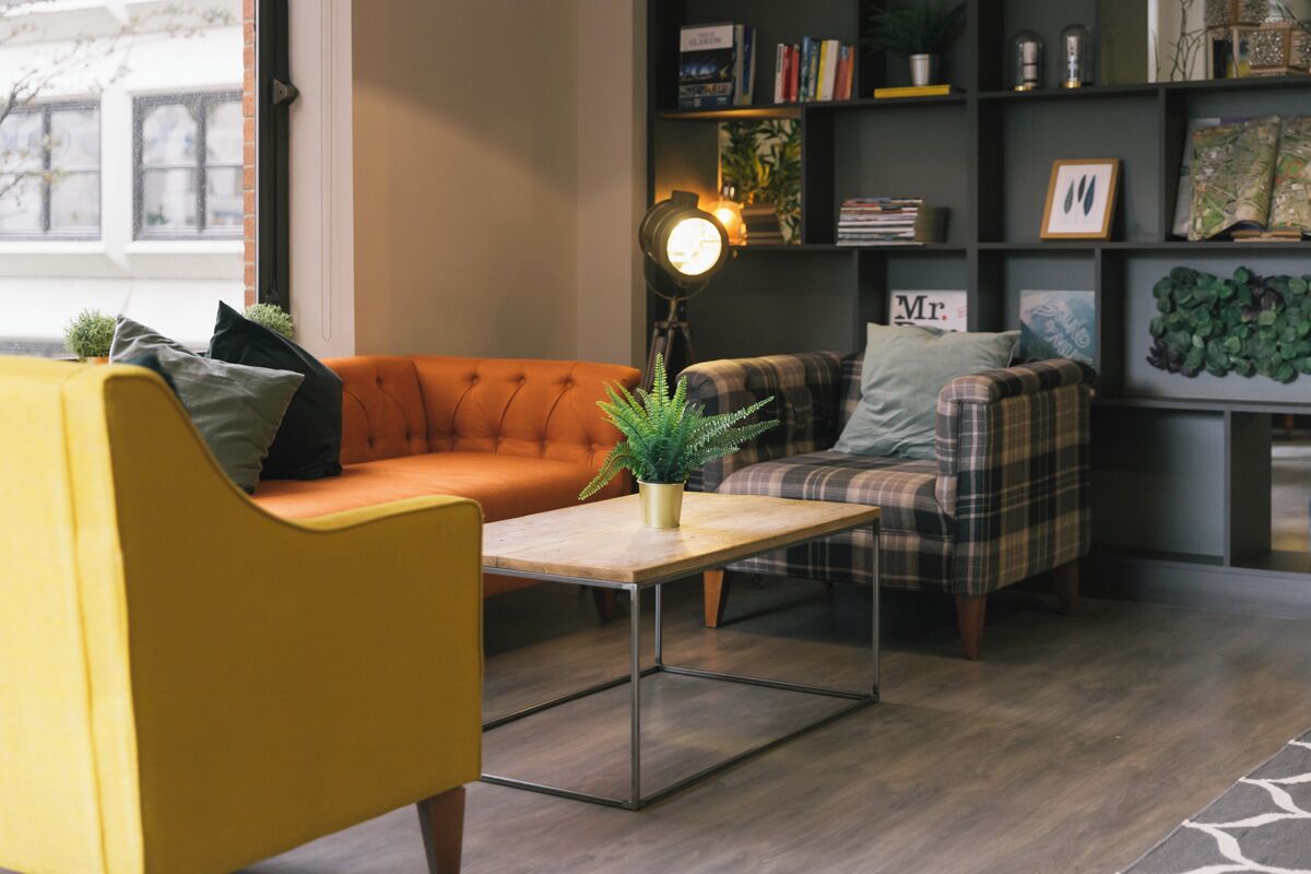 Una sala de estar acogedora con un sillón amarillo, un sofá naranja y un sillón a cuadros. Una mesa de centro de madera con una planta está en el centro. En el fondo, una estantería con libros y objetos decorativos.