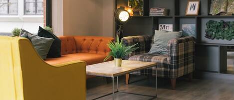 Una sala de estar acogedora con un sillón amarillo, un sofá naranja y un sillón a cuadros. Una mesa de centro de madera con una planta está en el centro. En el fondo, una estantería con libros y objetos decorativos.