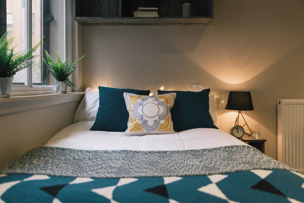 Ein ordentlich bezogenes Bett mit einem gemusterten Überwurf, ein Nachttisch mit einer Lampe und zwei Blument öpfe am Fensterbrett.