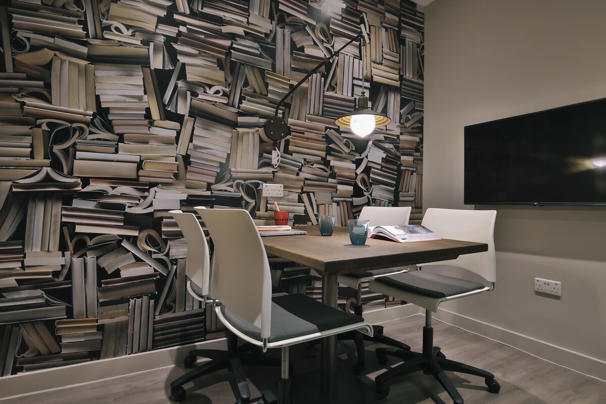 Un salón de conferencias con una pared repleta de libros, un escritorio con sillas, una lámpara y un televisor de pantalla plana.