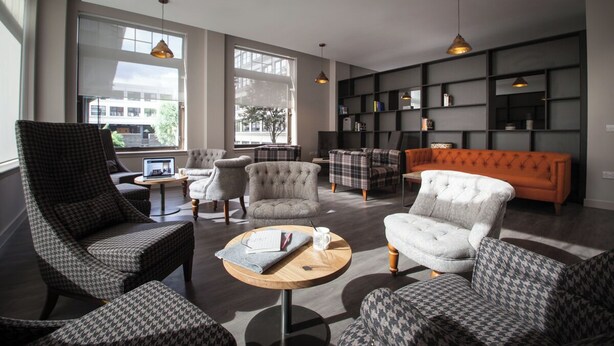 A modern living room with a sofa, armchairs, and a coffee table. There are bookshelves and a large window providing natural light. - Student Only Bath Street (Glasgow)