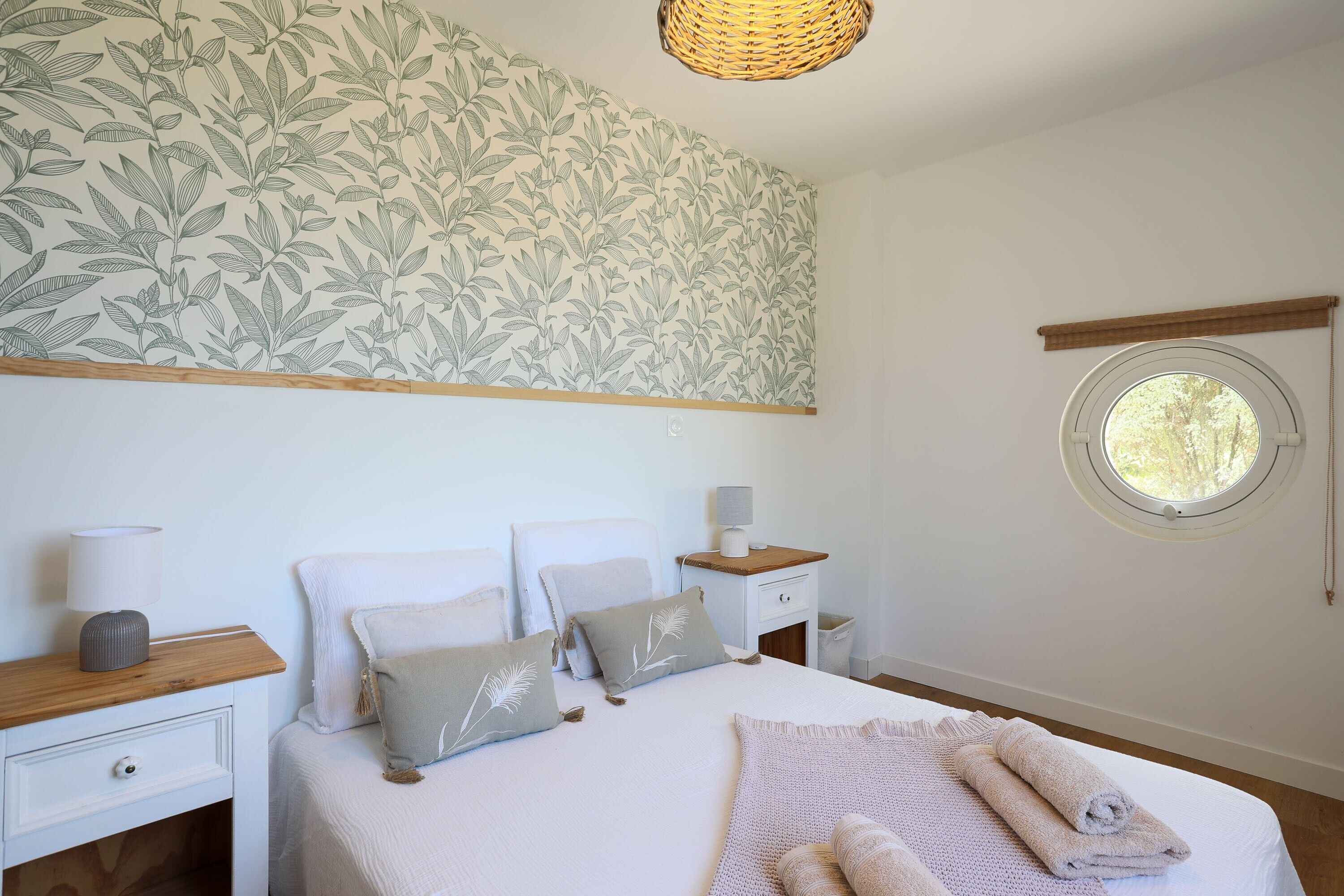 A bedroom with a bed, pillows, a nightstand, a lamp, a window, and a wall with a leaf pattern.