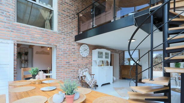 A modern interior with a wooden dining table, a spiral staircase, and a brick wall.