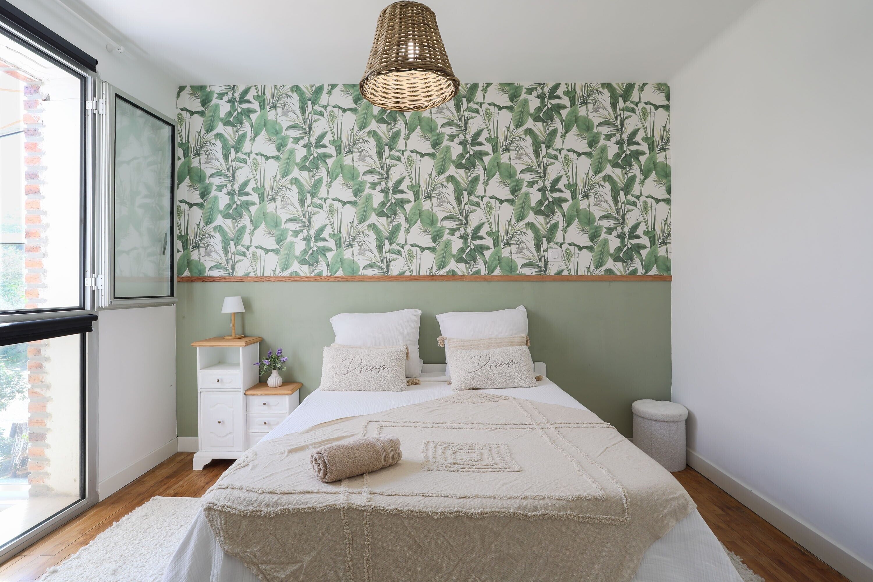 Un dormitorio con cama, mesita de noche, lámpara, ventana y pared con diseño de hojas.