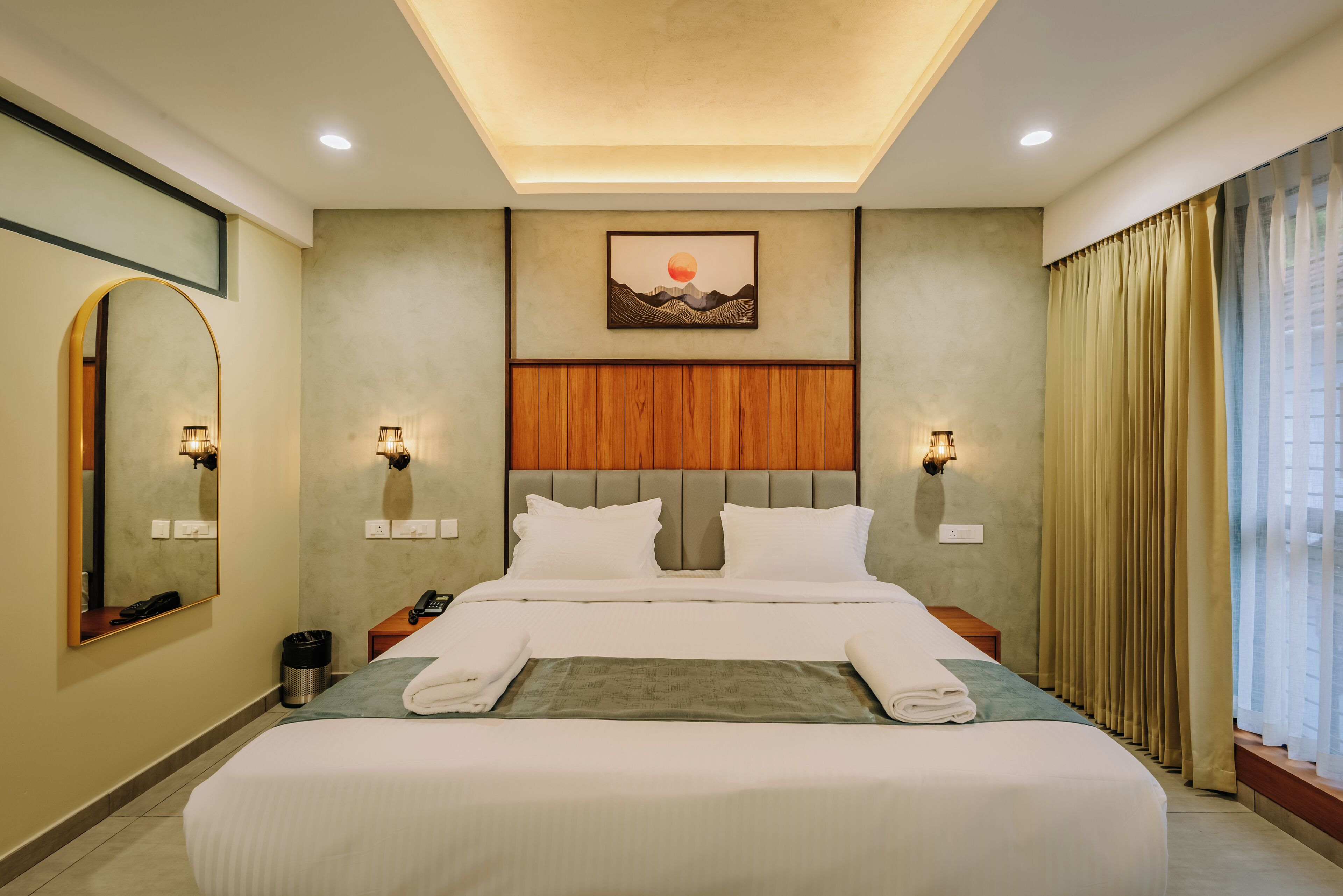 Une chambre d’hôtel avec un grand lit, une tête de lit en bois, deux tables de chevet avec des lampes, un miroir fixé au mur et un tableau représentant un coucher de soleil.