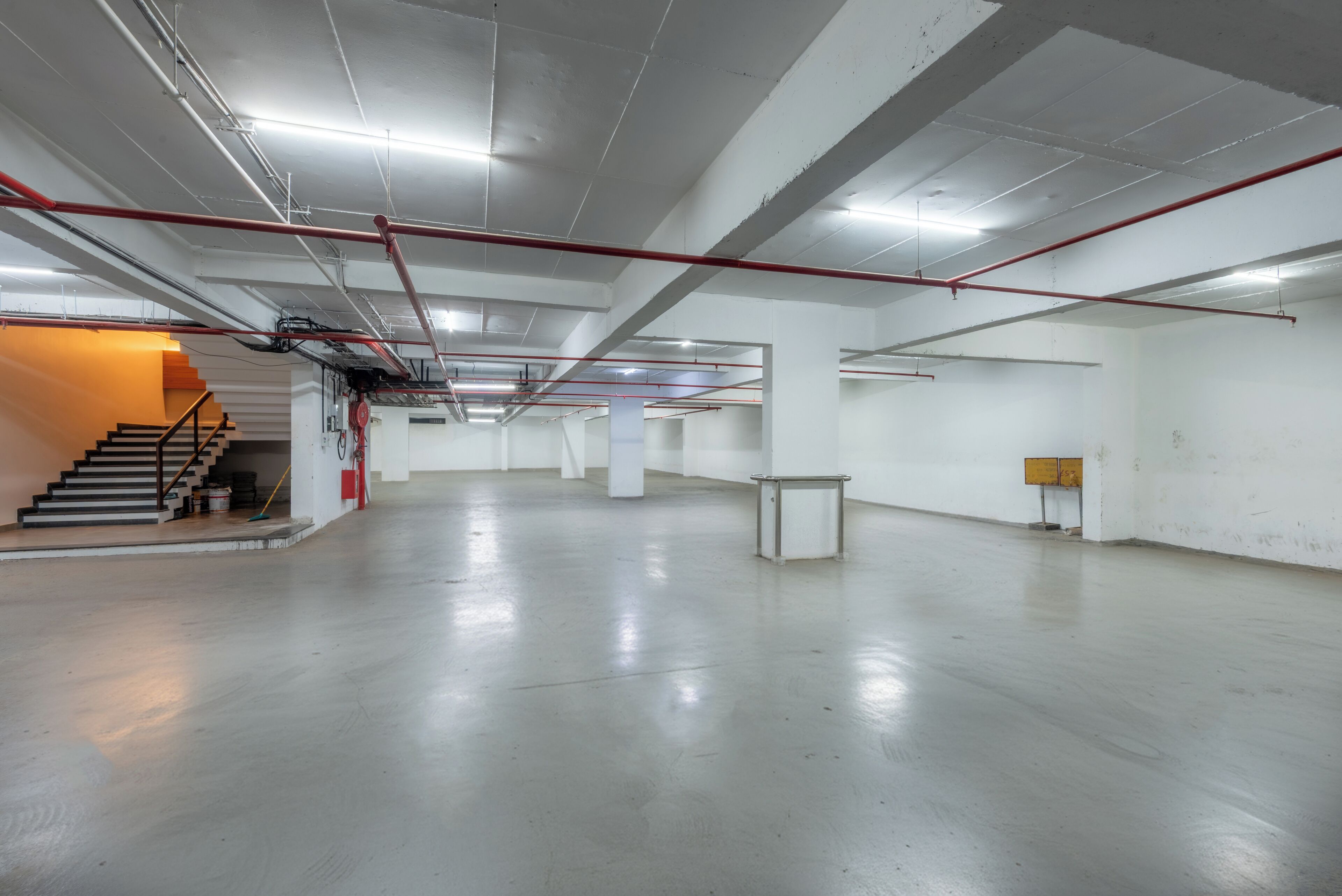 A spacious, empty room with a polished concrete floor, white walls, and a staircase on the left. Red pipes and fluorescent lighting are visible on the ceiling.