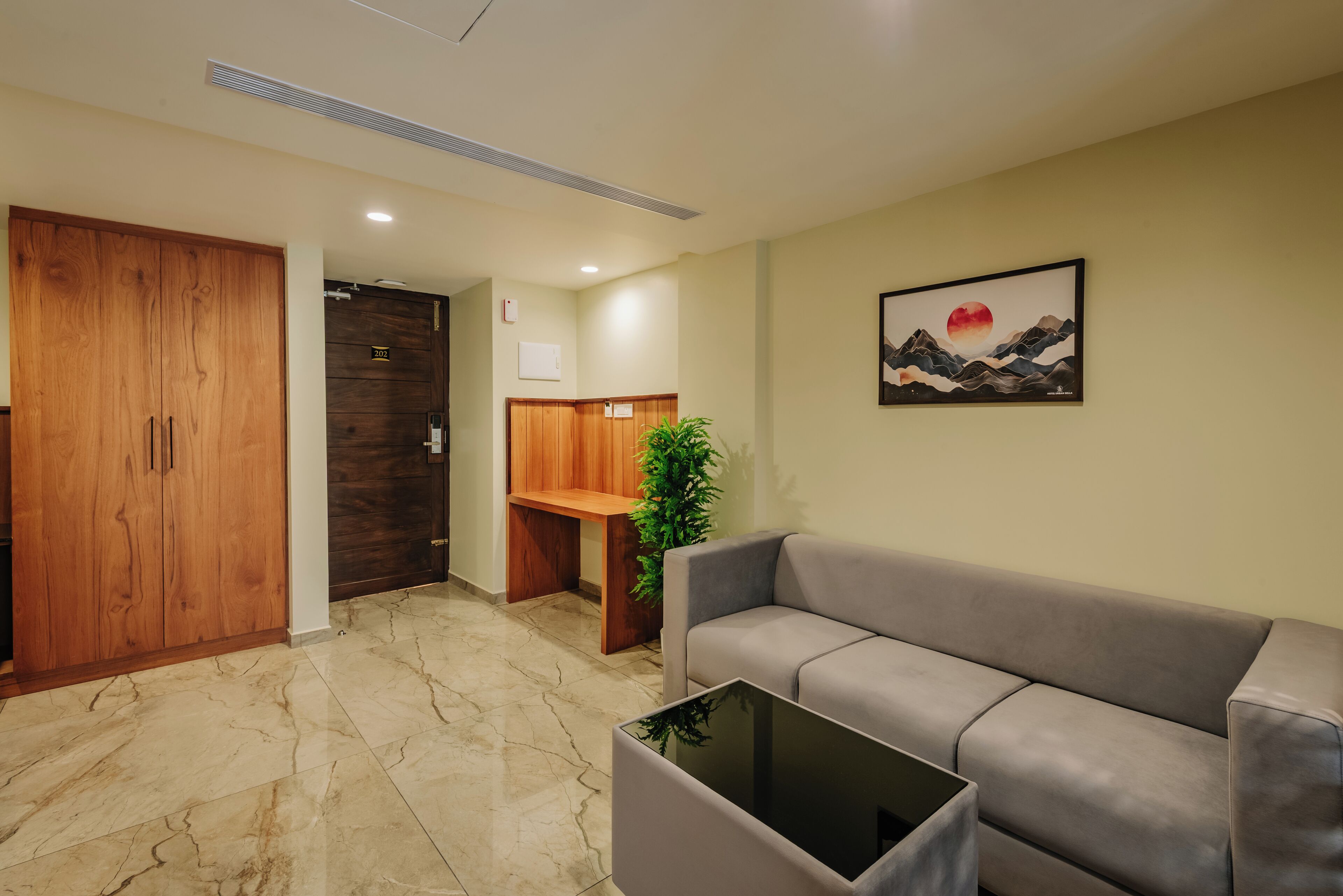 A hotel lobby with a wooden wardrobe, a reception desk, a grey sofa, a glass coffee table, and a framed artwork on the wall.