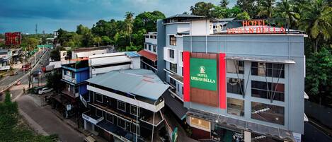 Un hotel de varios pisos con un letrero llamativo que dice 'HOTEL URBAN BELLA' y vistas del área urbana circundante, incluyendo calles y zonas verdes.