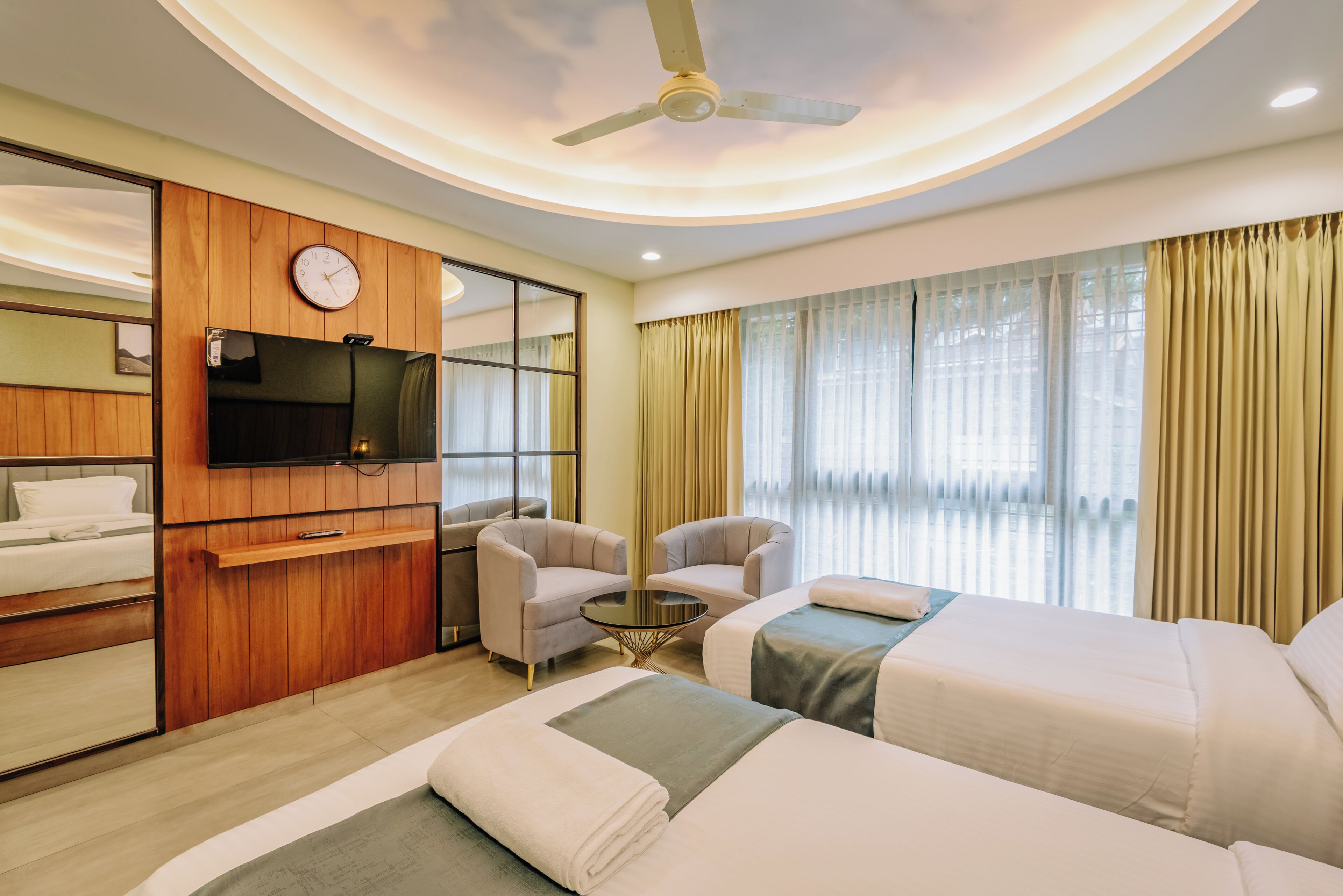 Habitación de hotel con cama, dos sillones, una mesita, televisión montada en la pared y ventilador de techo.