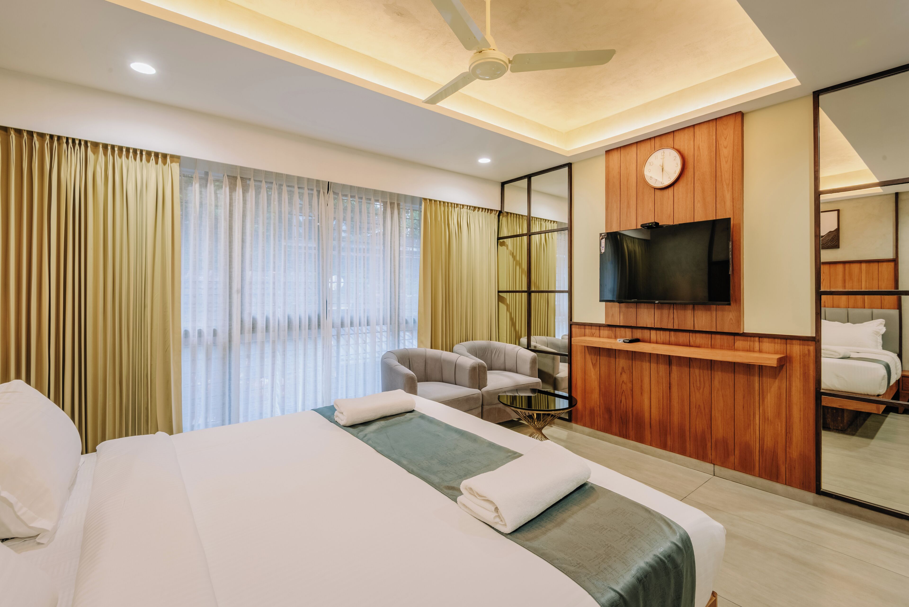 A hotel room with a large bed, a television mounted on the wall, a ceiling fan, and a window with curtains.
