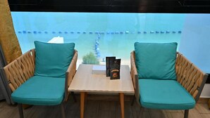 Two teal cushioned chairs with wooden legs and a small table in between, placed in front of a large window with a view of a blue sky and a distant road. - La Fontain lagoon resort  (Jeddah)