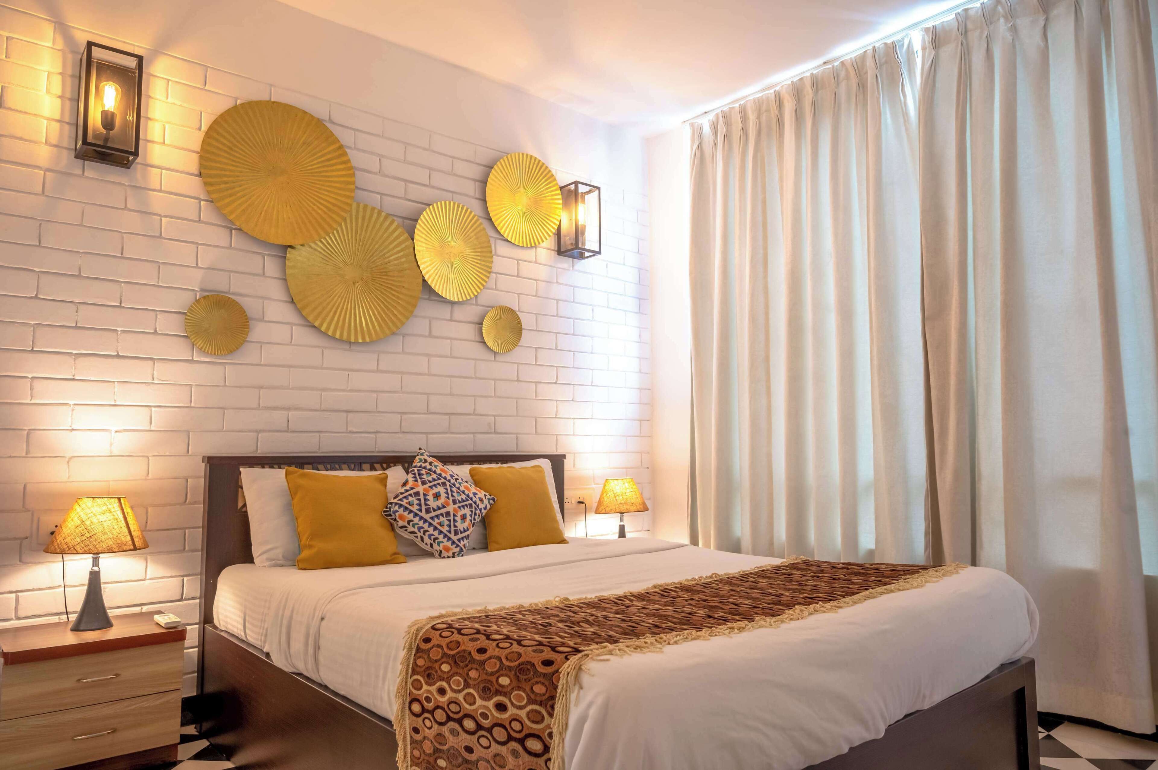 A hotel room with a white brick wall, a large bed with a patterned throw, two bedside lamps, and decorative wall art.