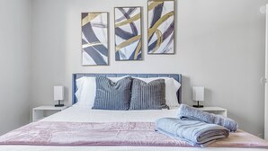 A neatly made bed with a white and pink patterned bedspread, two bedside lamps, and three abstract paintings on the wall.
