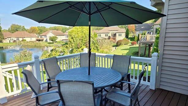 Een terras met een ronde tafel en stoelen, een parasol en uitzicht op een meer en huizen op de achtergrond.