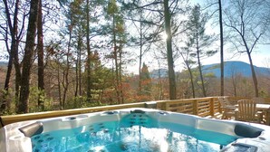 A hot tub with a wooden deck and chairs, surrounded by trees and a mountainous backdrop. - Luxury•5 Kings•MTN Views and Lake Access•FirePit•EVCharger•PoolTable (Lake Lure)