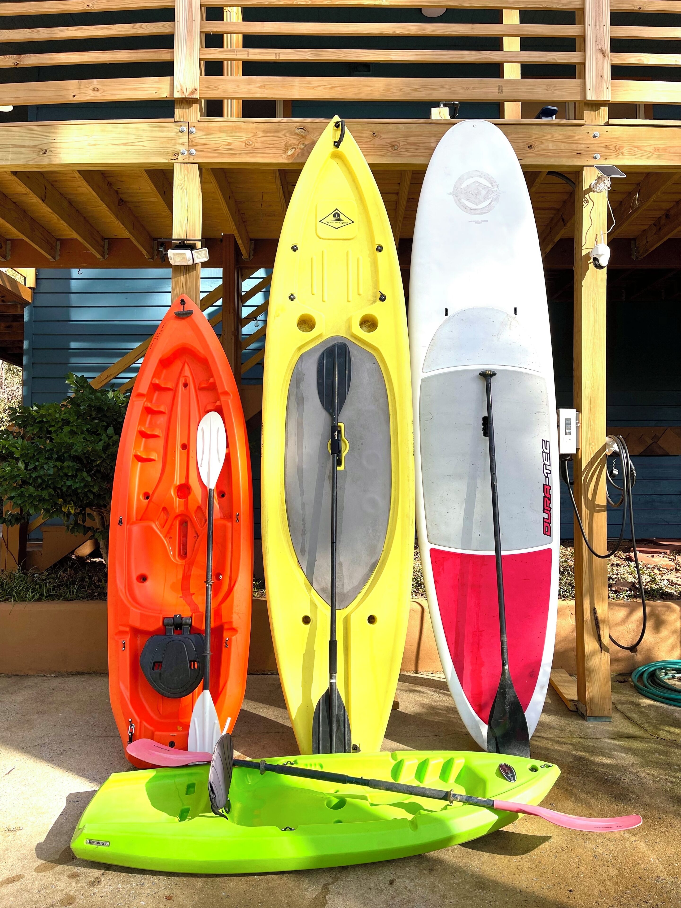 A row of kayaks and a paddleboard under a wooden structure.