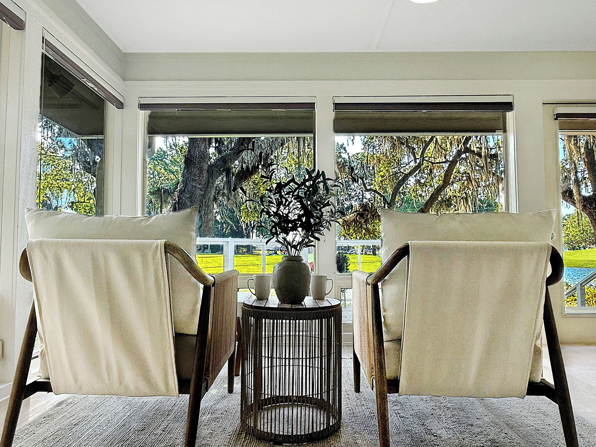 A room with two chairs facing a large window, a small round table with a vase and a cup, and a view of trees and a yellow fence outside.