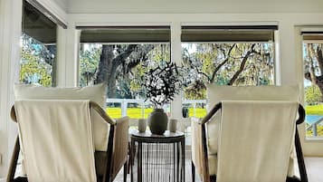 A room with two chairs facing a large window, a small round table with a vase and a cup, and a view of trees and a yellow fence outside.