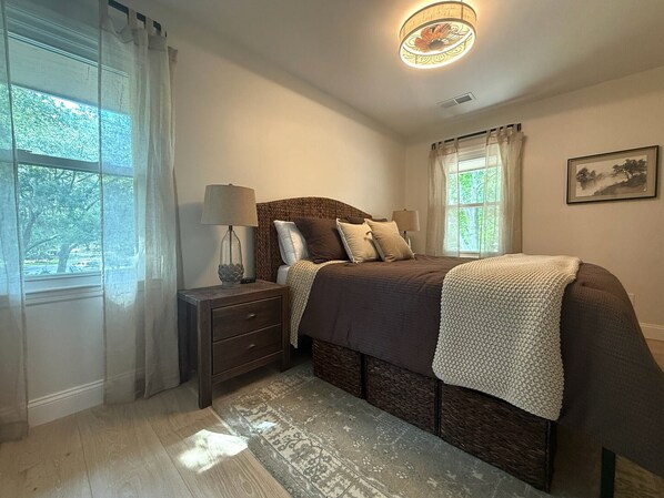 Un dormitorio con cama, mesitas de noche, lámpara, ventana con cortinas y un cuadro en la pared.