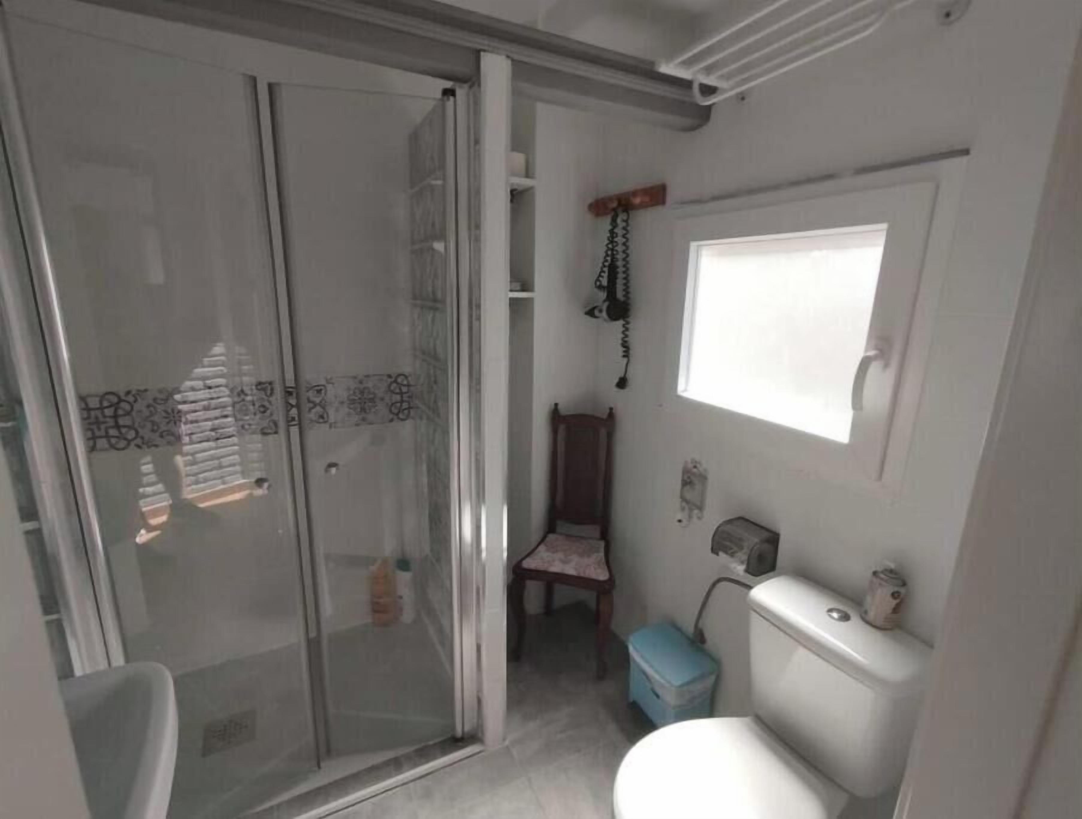 A bathroom with a glass shower enclosure, a toilet, a small window, and a wooden chair.
