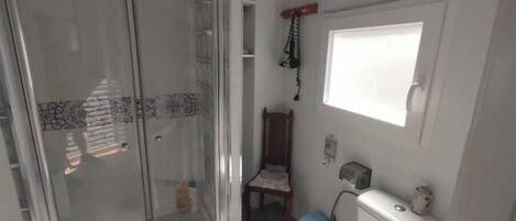 A bathroom with a glass shower enclosure, a toilet, a small window, and a wooden chair.