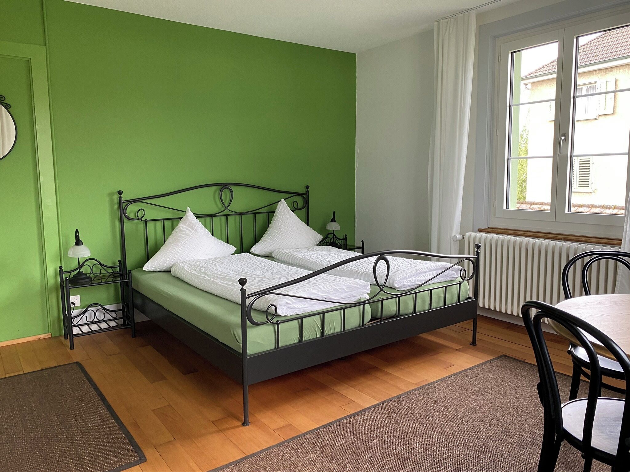 A room with a green wall, a metal bed frame, white bedding, a small bedside table with a lamp, a radiator, and a window with curtains.