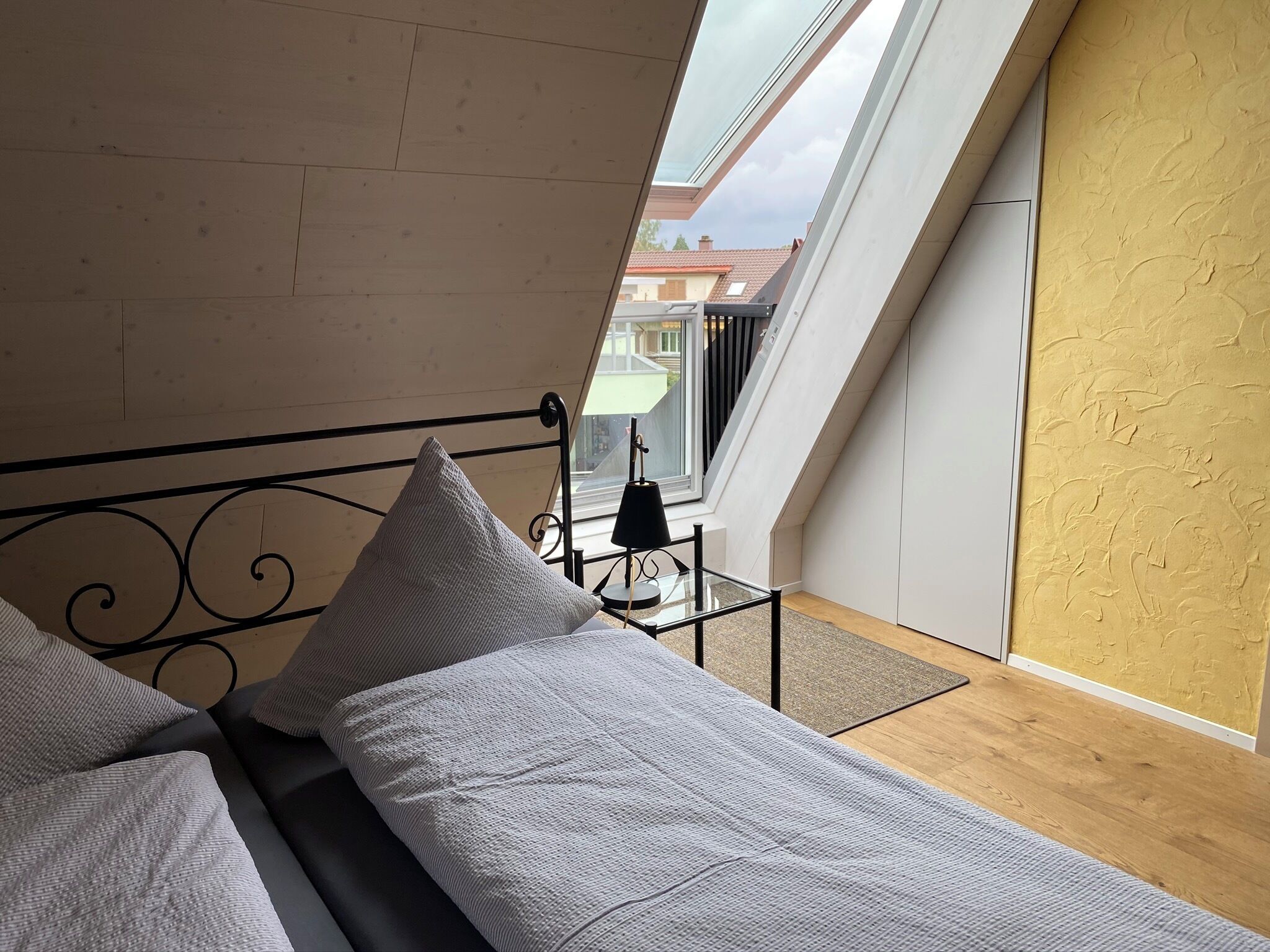 Ein Schlafzimmer mit Oberlicht, einem Bett mit Kissen, einem Nachttisch mit Lampe und Blick ins Freie.