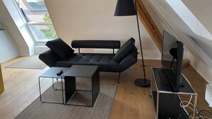 A modern loft bedroom with a sloped ceiling, a black sofa, a small table, a floor lamp, and a TV on a cart. - Apartment Hopfengrün Langenthal (Langenthal)