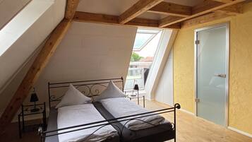 Une chambre avec un plafond en pente, un lit recouvert de draps blancs, une table de chevet avec une lampe, et une vue sur l’extérieur par une fenêtre.