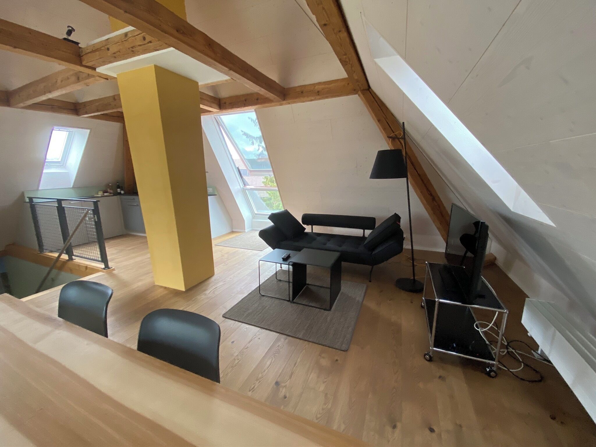 Une pièce de style loft avec un plafond haut, des poutres en bois, un coin repas avec une table et des chaises, un canapé, une lampe sur pied et un petit coin cuisine avec une fenêtre.