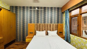 A room with a bed, wooden wardrobe, patterned wallpaper, and a window with curtains. - Hotel Indra (Srinagar)