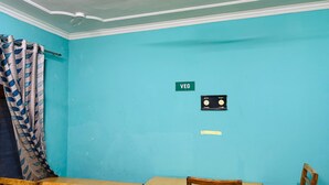 A room with teal walls, a wooden table, a chair, a curtain with a leaf pattern, and a wall-mounted electrical box labeled "VEG". - Hotel Indra (Srinagar)