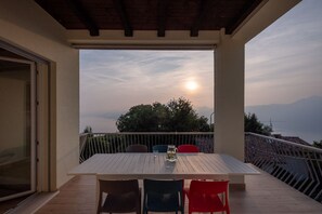 A balcony with a table and chairs, overlooking a scenic view of the sea and mountains.