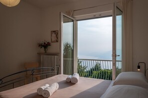Un dormitorio con cama, una silla, radiador, un florero con flores y un balcón con vistas.