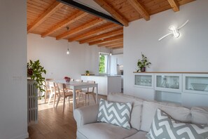 Ein modernes Interieur mit Holzboden, einem Essbereich mit Stühlen und Tisch, einer Küche mit weißer Arbeitsplatte und einer Sitzecke mit Sofa und Kissen.